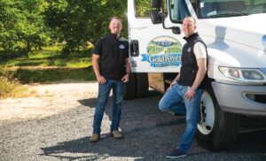 Two men standing in front of a white truck while smiling and looking at the camera.