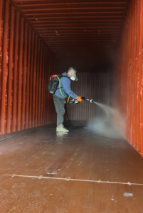 Man wearing protective face gear while spraying chemicals inside a shipping container.