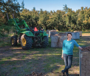 Woman standing in front of a large piece of farm equipment and bins full of hazelnuts, with trees in the background.