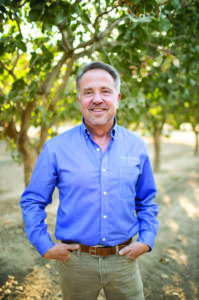 Man wearing a blue shirt and brown pants smiling while standing in front of pistachio trees.