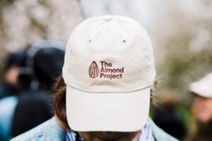 Person with a face covered by a white hat with the words The Almond Project in brown lettering.