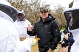 Man wearing a black jacket and camo hat surrounded by beekeepers in an almond orchard.