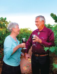 Couple smiling at each other while holding hands and glasses of wine and standing in a vineyard.