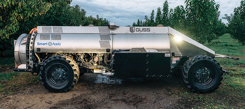 A large, silver piece of farm equipment parked in an orchard.