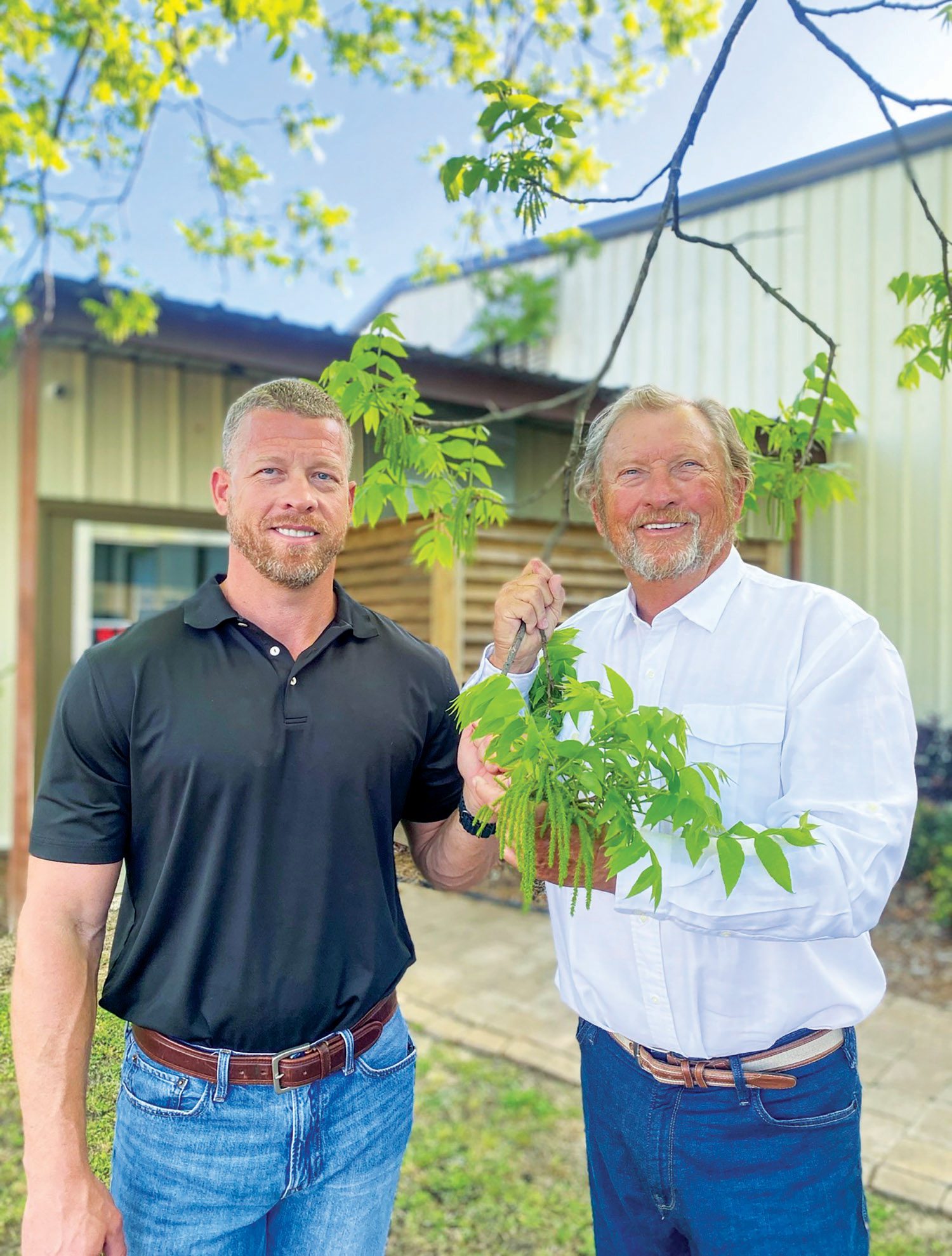 two men smiling while holding onto a pecan branch, as they are pecan farmers