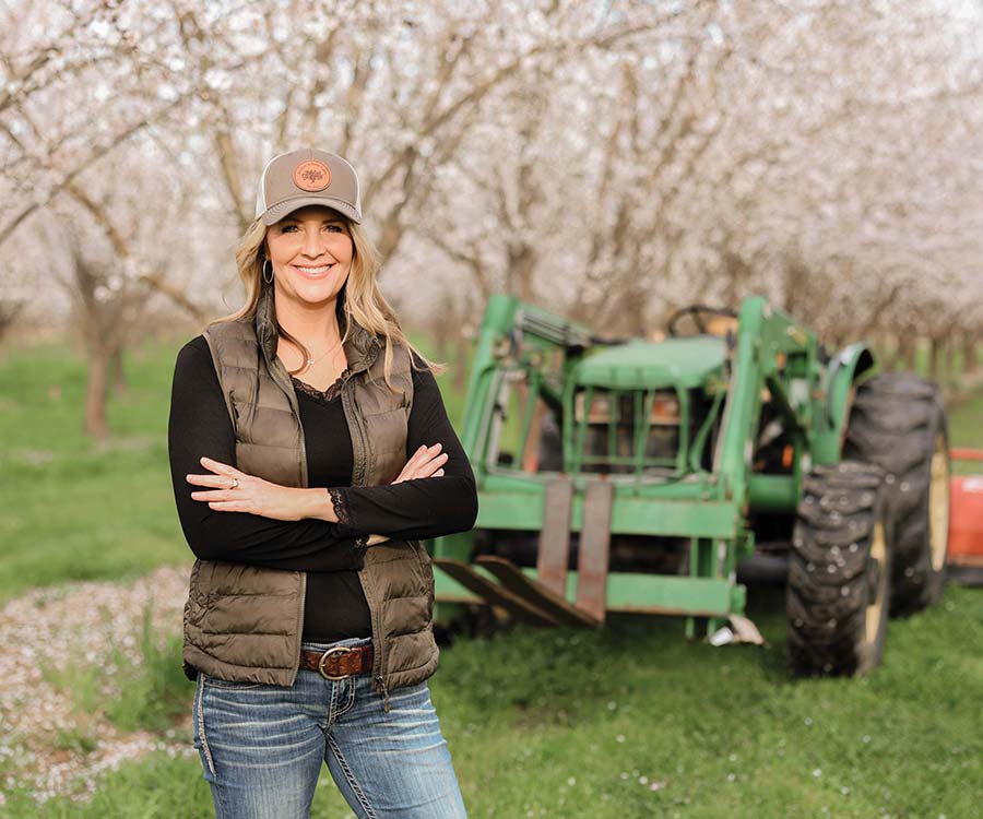 Woman standing in almond orchard with green tractor and trees in the background.